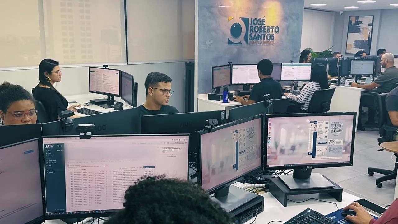 Sala de produção da JRS Cálculo Judicial: mais de dez pessoas em estações de monitor duplo, com a marca do escritório na parede ao fundo e as janelas da cidade à esquerda.