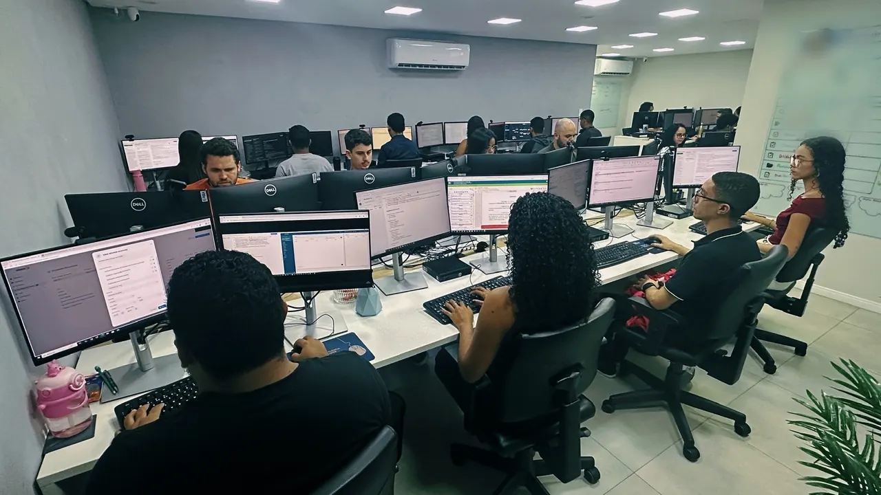 Vista ampla da sala de produção, com a equipe nas estações e o quadro da rota de produção na parede à direita.