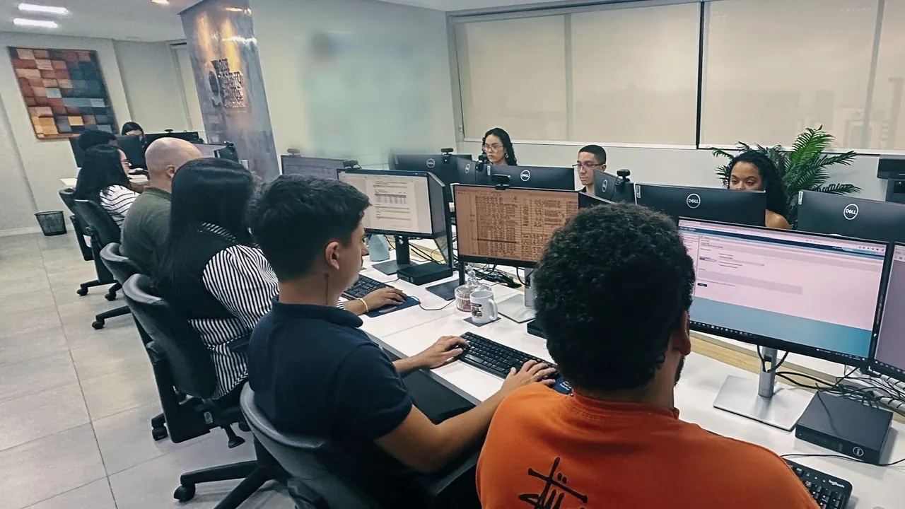 Plano fechado de uma fileira de estações, com a equipe de perfil diante dos monitores.